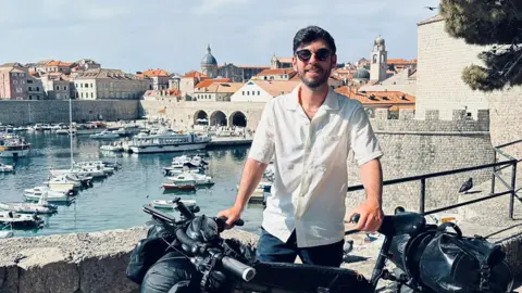 Nathan Hurley Man with dark hair and sunglasses poses with his bike in front of a harbour scene with boats and old stones buildings.
