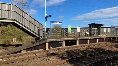Daniel Hodgson Platform two at South Bank is a short concrete platform with a waiting shelter and various signs erected. The steel footbridge on the left leads down to it. There are rail tracks in the foreground.
