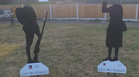 Michael Keohan/BBC Image shows a Tommy silhouette and a Women in War silhouette neither of which have heads. They are on a lawn in front of a fence
