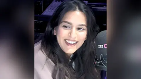 A woman with long, dark hair smiles as she talks into a microphone