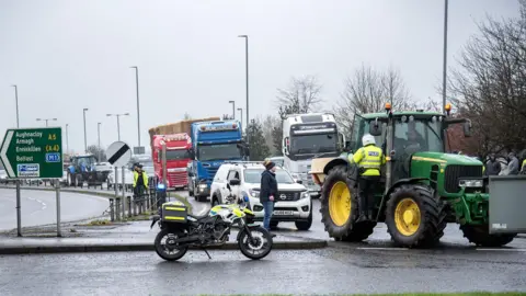 Pacemaker A number of tractors and trucks driving at a roundabout on a grey overcast day. A police motorbike is parked, where the police officer has gone to the tractor at the head of the convoy to speak to the driver.