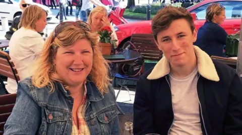 Two people - a mother and son - in an outdoor restaurant/cafe. The mother is on the left of the photo wearing a denim jacket and has blonde hair. The son has a dark blue jacket over a grey t-shirt and has neatly styled brown hair. 