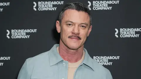 Headshot of Luke Evans. He wears a light blue jacket and cream jumper. He has greying hair and a moustache 