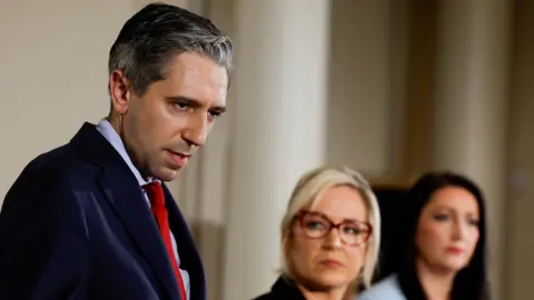 Simon Harris speaking to media - he has brown hair and wearing a navy suit with a light blue shirt and red tie, he is standing beside Michelle O'Neill, a blond haired woman wearing red glasses and Emma Little-Pengelly, a long brown haired woman. 