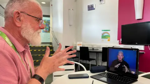 A man sits at a table indoors, gesturing with their hands while taking part in a video call on a laptop.
