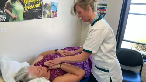 Nikki Fox/BBC Occupational Therapist Sara Heritage stands over Deborah Caitens-Smith who is lying on a medical bed. Ms Heritage is wearing white scrubs with a dark green edging and she has blonde tied back hair. She is feeling the chest of Ms Caitens-Smith who is looking at her.
