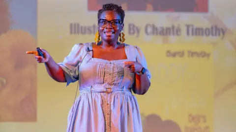 A woman with short curly hair stands on stage, seemingly presenting. She is wearing a light blue dress and black glasses with thick frames. The background behind her is a bright yellow projection.