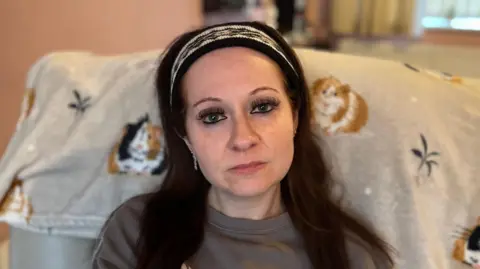BBC A woman with black hair with a white head band on stares directly into the camera. She has long eyelashes and a brown top on. She is sat on a chair which has images of guinea pigs printed on it. 