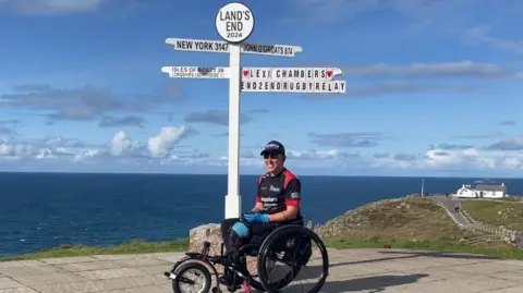 BBC Ms Chambers, wearing a black and read sports shirt, in her wheelchair. Behind her is the lands end sign pointing at New York, John O'Groats and Isles of Scilly. There is one sign that says Lexi Chambers END2ENDRUGBYRELAY on it. The background is the sea. In the left of the background is a hill with a white house. 