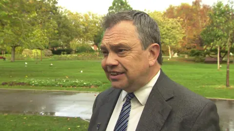 Picture taken in Victoria Park, Portsmouth. Trees are visible in the background. Cllr Sanders is wearing a grey jacket with white shirt and blue striped tie
