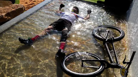 Javier Martinez-Zubiko Rolls lies in a small pool of water along with his bike. He is still wearing his bike helmet and clothes as he smiles.