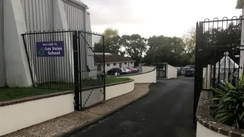BBC The gates to enter the Les Voies school are open, black and and there is a small sign to the left reading: Welcome to Les Voies School.
