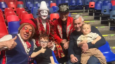A man and two young children sit with three performers from the circus. One performer, dressed as a clown has a red nose, a red hat and is wearing a blue waistcoat. Another performer is wearing a red costume with a white face paint while a third performer is dressed as the ringmaster in a red suit, black shirt and a black hat with gold detailing.