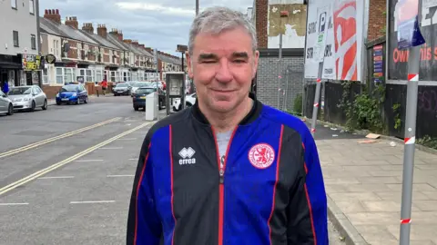 Robert Nichols standing on Linthorpe Road. He has short grey hair, brushed to his right, and is wearing a blue-and-black Middlesbrough FC tracksuit top. A row of terraced houses can be seen in the background on the left, while on the right are two advertising billboards.