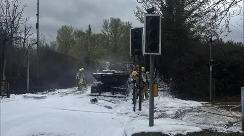 Wellington Fire Station A road is covered in white foam, with a team of firefighters wearing yellow helmets surrounding a burned-out vehicle in the centre of the image. There are traffic lights in the foreground, which are not lit. In the distance are trees and hedgerows.