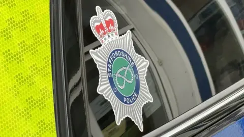 BBC A Staffordshire Police emblem on the window of a police vehicle which features a crown and a Staffordshire Knot. There is a yellow high visibility section of the vehicle on the left hand side of the image.
