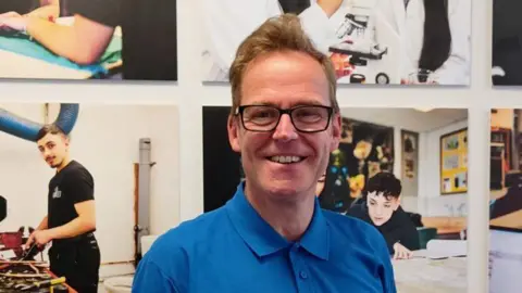 Keighley College A man in a blue t-shirt with black glasses stands in front of a poster showing teenage boys learning building skills 