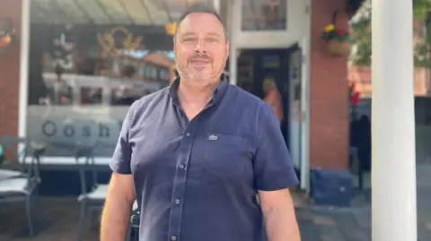 BBC/Angela Ferguson A man with short grey hair and a beard is smiling while standing outside a cafe. He is wearing a dark blue shirt. A hanging basket of flowers can be seen blurred out behind him, along with part of a window sign saying 'Ooshka Ba'.