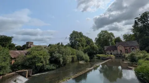 BBC Weather Watchers / George Groundhog A river leads up to a brick house with a series of large green trees around it which are reflected in the river.