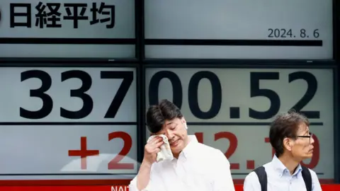 Reuters Two men in front of an electronic board displaying the Nikkei stock average outside a brokerage in Tokyo, Japan, 6 August, 2024.