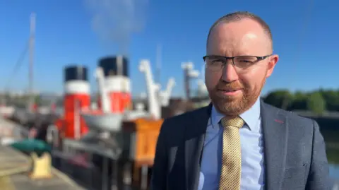 Paul Semple, a bearded man in a grey suit and gold tie, smiles with the Waverley out of focus behind him.