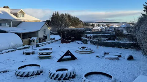 Snow has settled on a school playground. It is covering a number of playground furniture including outdoor benches and tractor tyres which have been placed on the ground. Buildings in the distance are also covered in snow. 
