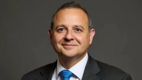 UK Parliament Head and shoulders shot of man in a grey suit and blue tie.