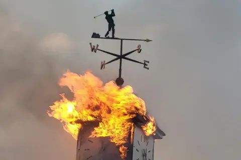 Ferndown Fire Station Rood alight with square clock tower with weathervane golfer silhouette on the top - flames are coming out from the sides of the clock.