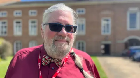 Michael Fife Cook wearing dark glasses and a burgundy shirt and a bow tie and a lanyard