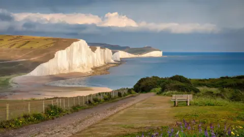 Picturesque white cliffs, known as the Seven Sisters, face a pale blue ocean. The sun is setting. 