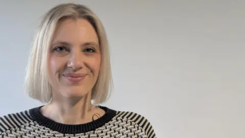 Head and shoulders of a woman with shoulder length short blonde hair, standing in front of a white backdrop.