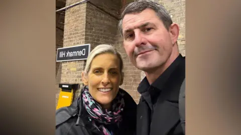 A picture of Lisa and her husband Justin. They are stood in a train station with a brick wall behind them.