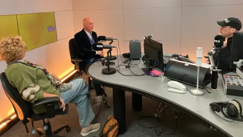 The image shows a radio studio. Sitting in the foreground is Kim Holloway, wearing a green cardigan, jeans and beige trainers. Behind her is Ian Holloway sitting in a dark suit talking to the radio presenter Joe Sims. He wears a dark top, glasses and a baseball cap. He is sitting next to a microphone, behind the desk in the studio.