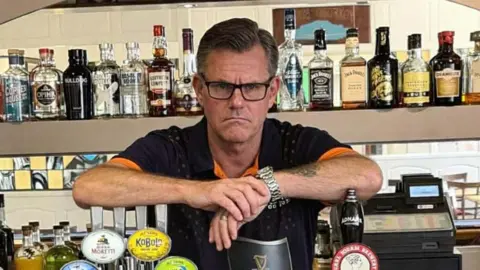Supplied An angry looking Dennis Wilson. He is standing behind a bar with his arms resting on some beer pumps. He is wearing glasses and a black-and-orange top while looking into the camera. 