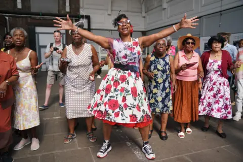 PA Media Women in colourful dresses dance at a reception following the arrival of a Thames Clipper at the Port Of Tilbury with 100 NHS workers and 100 people with Windrush connections to mark the 75th anniversary of the arrival of the Empire Windrush. Picture date: Thursday June 22, 2023.