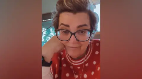 Maria Elliott A woman with short grey hair and glasses smiles at the camera, resting her head on her chin. She wears a red jumper with white spots. 