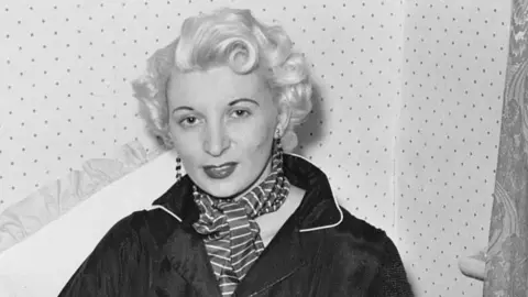 Ruth Ellis in black and white photo taken before her arrest wearing striped scraf and dark collared jacket, long earrings and she has short blonde hair. 