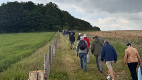A long line of people walking up a wide path through fields. There are large trees ahead of them and a fence either side of the path.