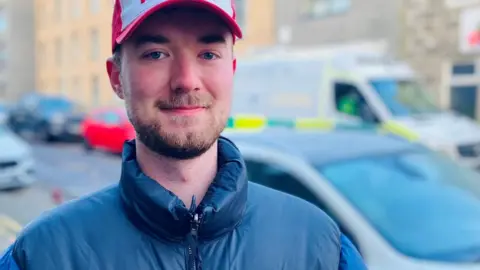 Patrick Alexander, man smiling and looking at camera, wearing a warm blue jacket, red and white cap, on a street with cars and ambulances in background.