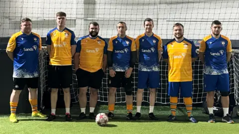 Family handout A football team lines up for a photo in front of a football net. There are seven men, wearing blue and yellow kit. They are stood on green grass and a football is in front of them.