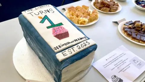 Kingsthorpe Community Library A cake in the shape of a blue book with writing on top. In the background are other small cakes. They have been placed on a table.