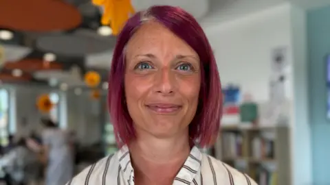 A head and shoulders photograph of a woman with hair dyed dark pink in a bob. She is wearing a white blouse with red stripes.