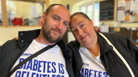 On the left Scott has receding, short dark brown hair and a goatee beard. On the right, Tina has mid-length brown hair which is tied back in a tight ponytail. The couple are leaning towards each other and touching their heads together. Both are wearing a diabetes charity t-shirt under black coats. They are smiling.