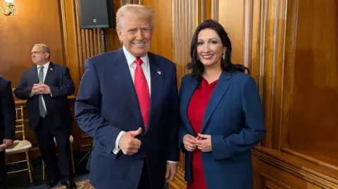 Donald Trump gives a thumbs up as he meets Deputy First Minister Emma Little-Pengelly. She is wearing a red dress with a blue blazer. The pair are standing in a wooden panelled room.