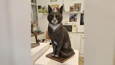 Tracey Jones A life-sized statue of a black and white cat sitting on a metal base. The statue is in an art studio and on the wall behind it are photographs of other statues.