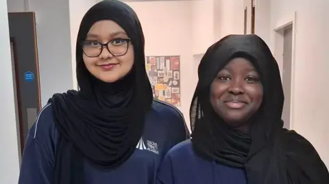 BBC Two girls stand alongside each other in a school corridor. They are wearing blue sweatshirts that have the Dixons Trinity Academy logo on them. The girl on the left is wearing glasses and a black headscarf. The girl on the right is also wearing a black headscarf.