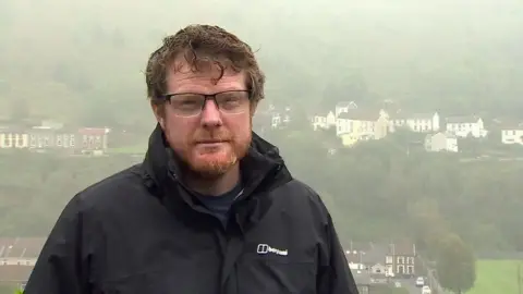 Phil Thomas of Clear South Wales' Coal Tips campaign group standing on a hillside with house behind him across a valley