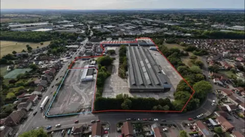 An aerial view of the former Hotpoint factory, a huge rectangular grey building with lines of angled roofs. The site is surrounded by grey car parks and a ring of green trees, with houses beyond. 