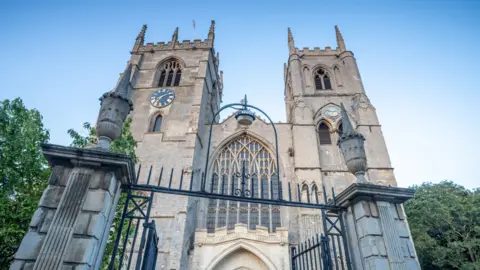 Getty Images Image from the ground looking up of a historic church with two towers and an ornate gateway. The left-hand tower has a clock face on it.