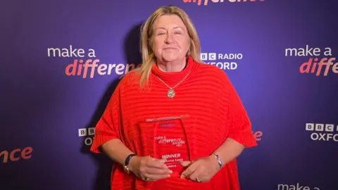 A smiling Grace Muir with her Make A Difference Animal award. She is standing in front of a purple Make A Difference branded wall.
